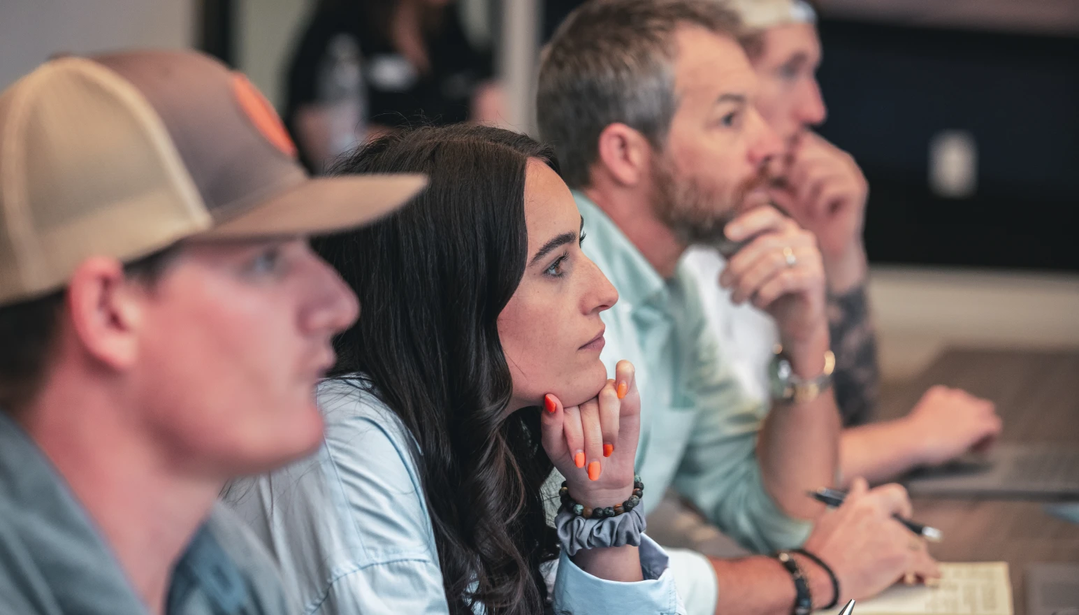 Four people, three men and one woman, sit attentively at a table, pens in hand, gazing forward. The mood is focused and contemplative.