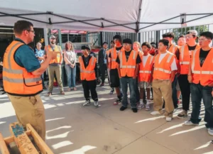A group of students in orange safety vests attentively listen to an instructor under a canopy tent. The setting conveys a tone of focused learning.