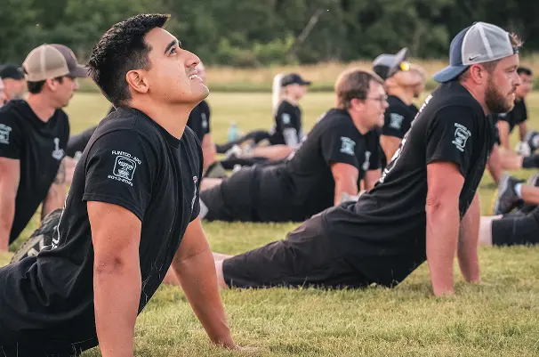 A group of people outdoors doing a yoga pose on grass, wearing matching black shirts. The setting is serene with a focus on a man smiling upward.