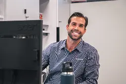 Smiling man in a checked shirt sits at a desk beside a monitor, creating a welcoming atmosphere. A water bottle is visible in front, emphasizing a casual workspace.