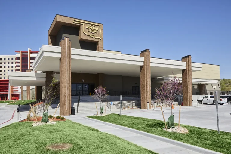 Front view of a modern building with a Hard Rock Café sign, featuring large columns and a beige facade. Surrounded by green lawns under a clear blue sky.