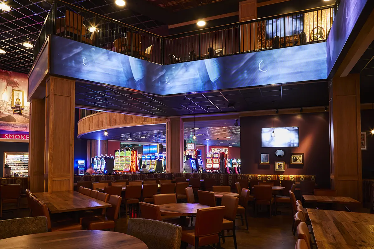 Spacious casino interior with warm lighting, featuring rows of colorful slot machines, round wooden tables, and comfortable chairs. It conveys a lively, inviting atmosphere.