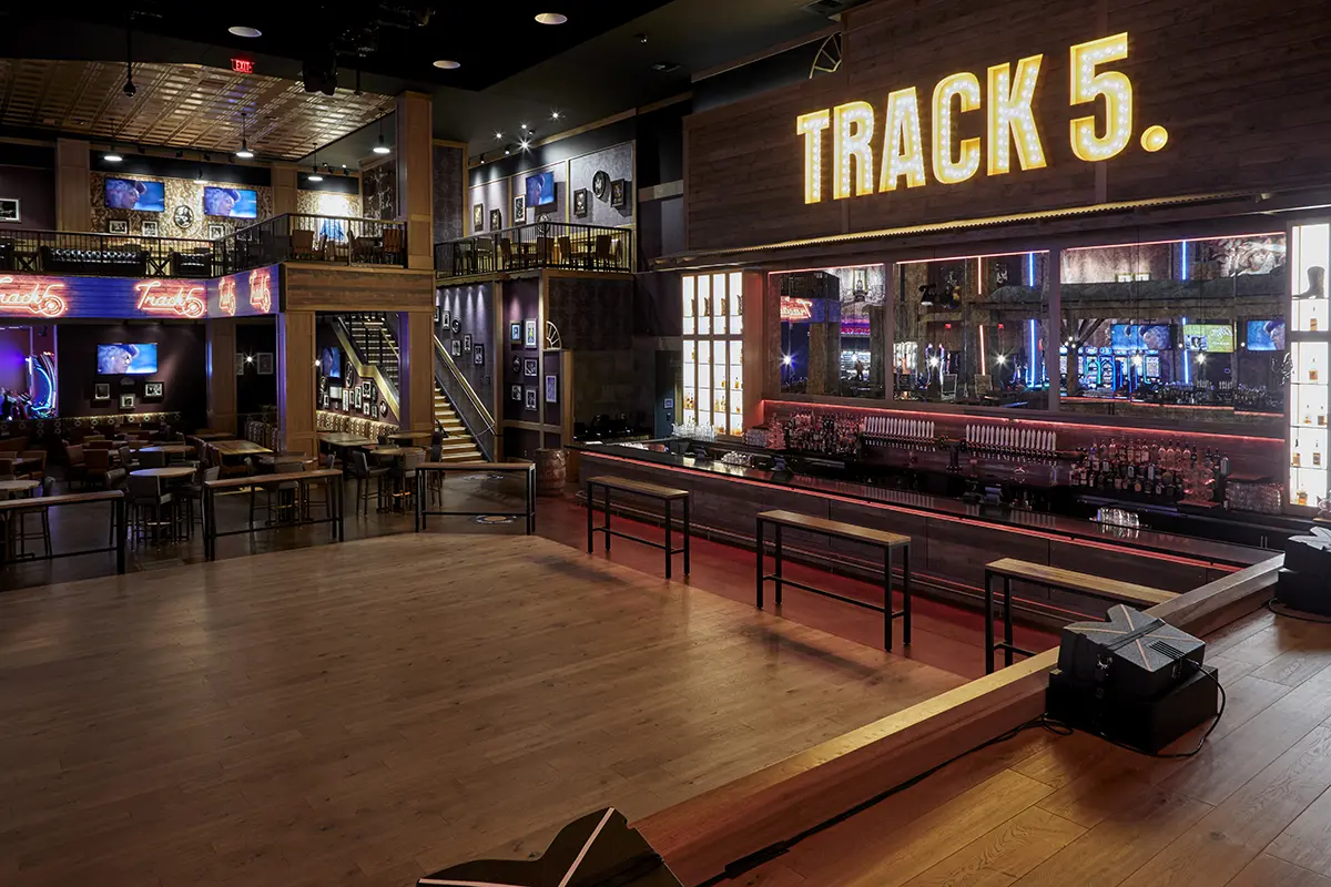 Dimly lit bar named “Track 5” features a spacious wooden dance floor, neon signs, and a long bar with stools. Upstairs seating adds a cozy vibe.
