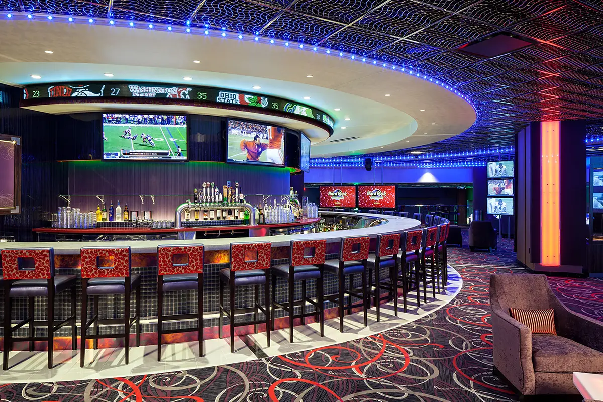 Vibrant sports bar interior with a curved bar, red-patterned stools, multiple TVs showing games, and blue ambient lighting, creating an energetic atmosphere.