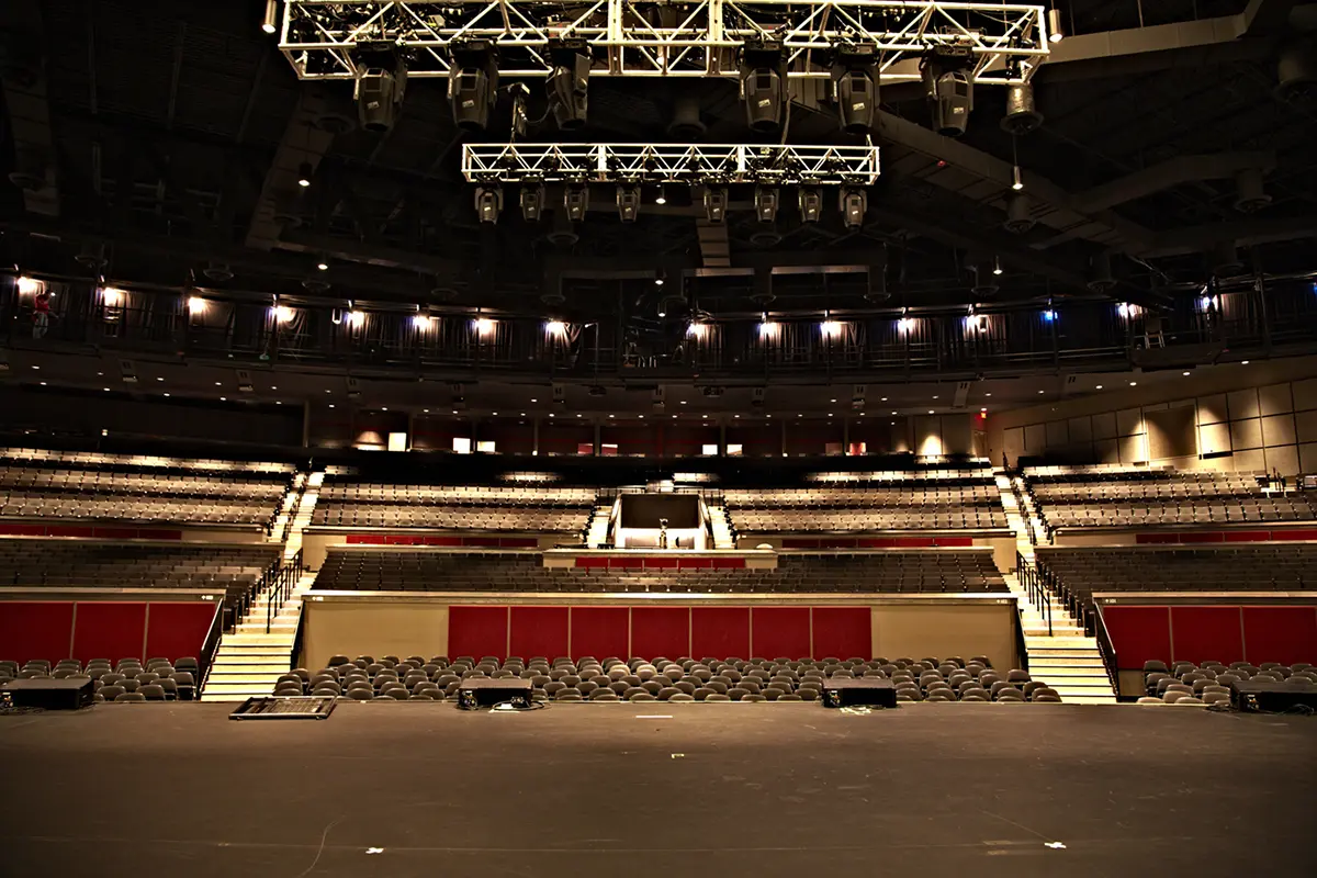 Empty theater auditorium viewed from the stage, featuring rows of seats, dim overhead lighting, and a calm, anticipatory atmosphere.