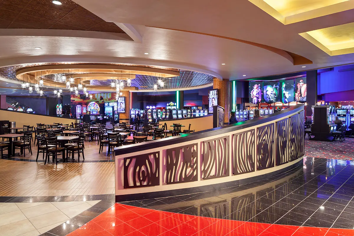 Spacious casino interior with glowing slot machines on the right, a circular area with empty tables and chairs on the left, and modern lighting above.