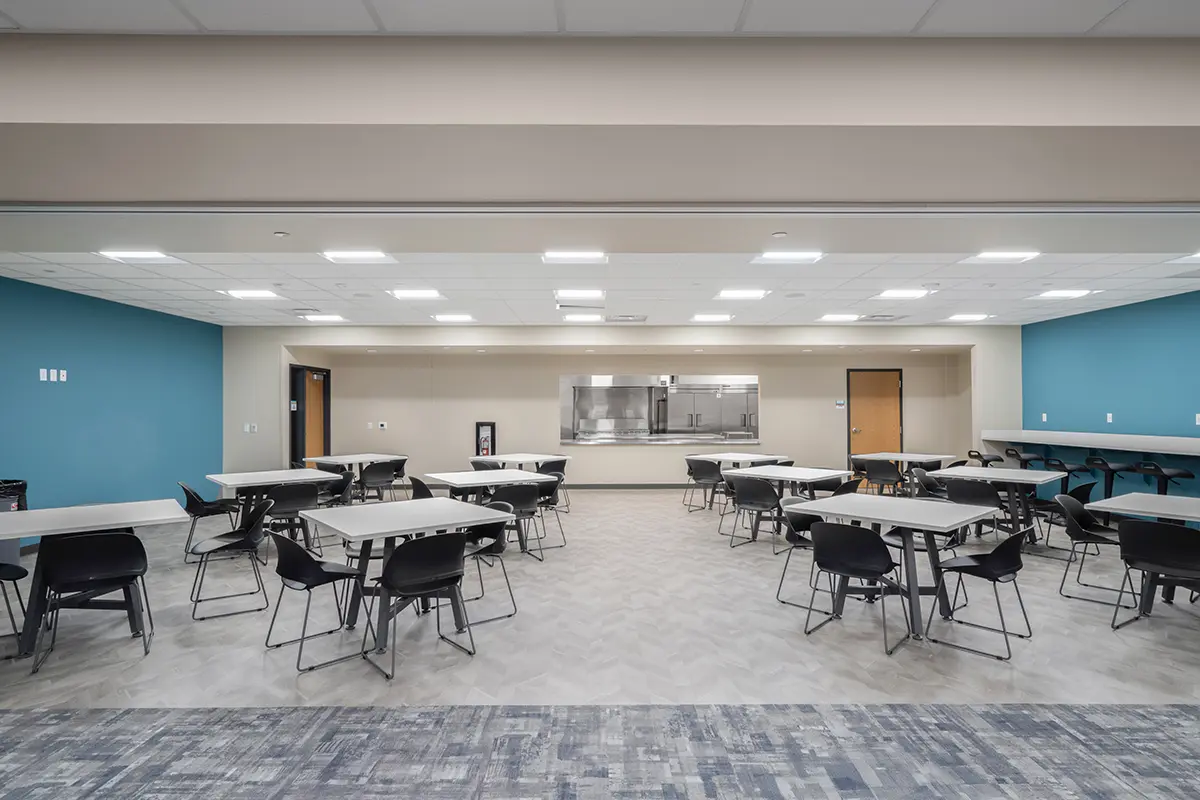 Spacious cafeteria with teal accent walls, rows of modern tables and chairs, and bright overhead lighting. Neutral tones create a clean, welcoming vibe.