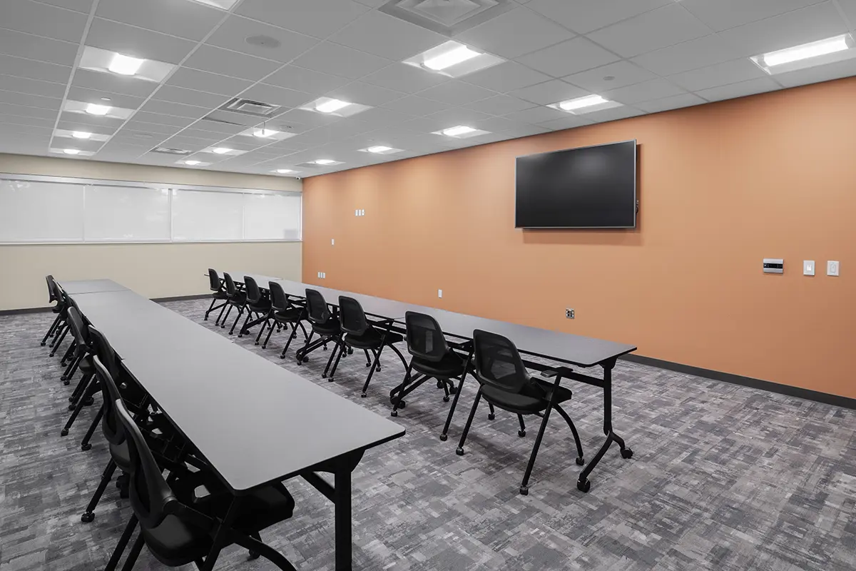 Modern conference room with long tables and black chairs arranged in rows. An orange accent wall features a large TV screen, exuding a professional tone.