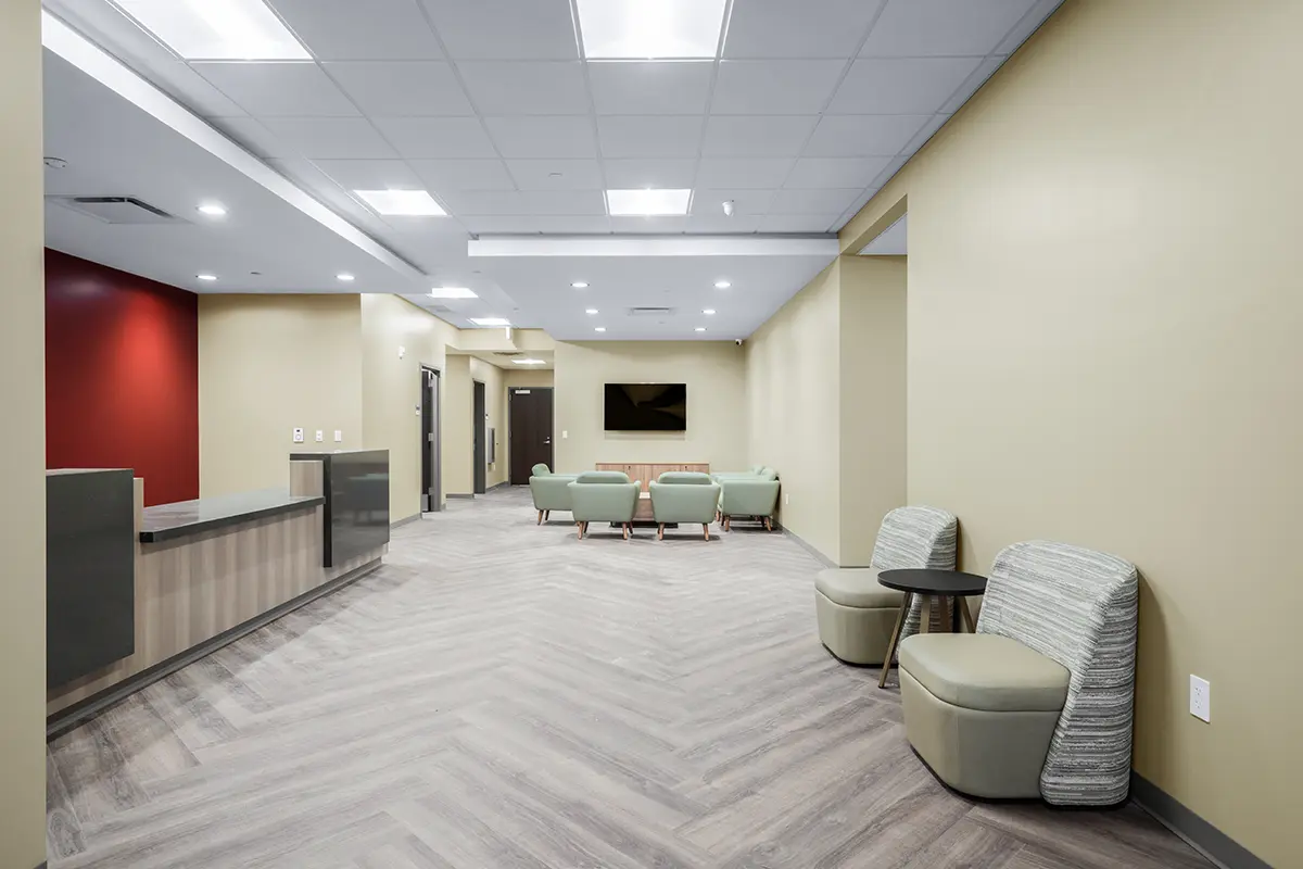 A modern waiting room with beige walls and wood flooring. Green chairs are arranged with a central TV on the far wall. The atmosphere is calm and inviting.