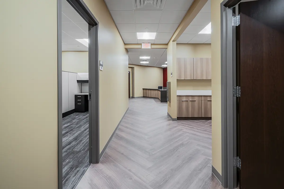 A modern office hallway with yellow walls, gray doors, and wood-patterned flooring. The space feels clean and organized, with cabinets on the right and a workspace visible on the left.