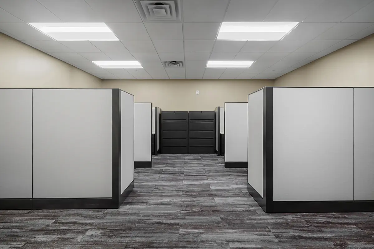 Empty office space with rows of beige cubicles under bright fluorescent lighting. Carpeted floor and neutral tones create a formal, quiet atmosphere.
