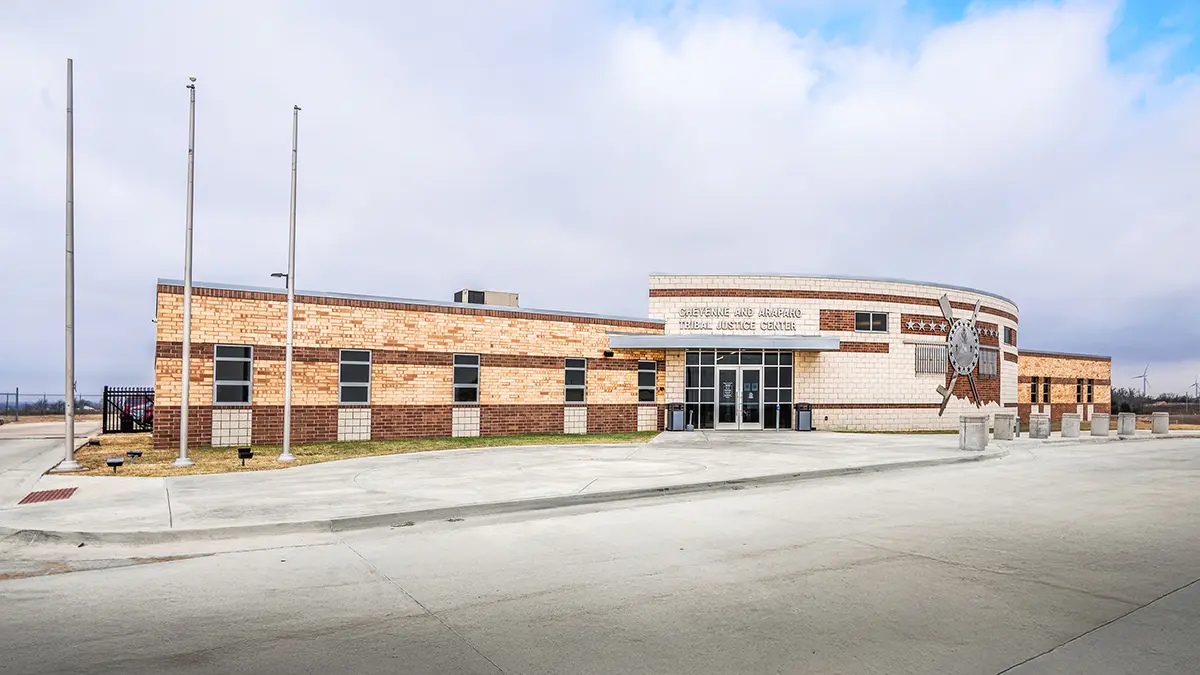 Large brick building with a curved facade, labeled "Detention and Judicial Complex" featuring tall flagpoles and a wide, empty pavement under a cloudy sky.