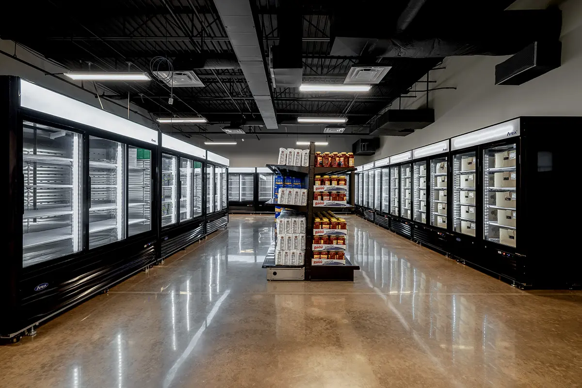 A brightly lit supermarket aisle displaying refrigerated cases on both sides with metal shelves. Central shelves hold bottled beverages and snacks.