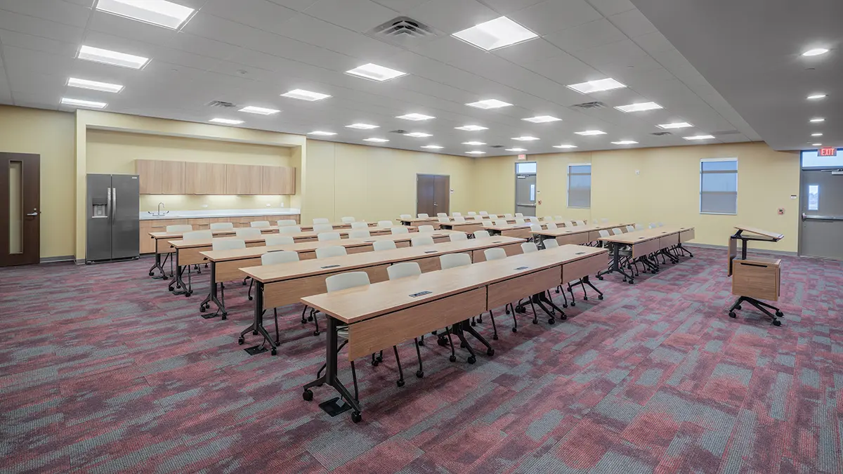 A modern classroom with rows of long wooden tables and beige chairs on a red carpeted floor, lit by bright ceiling lights and a cheerful yellow wall.
