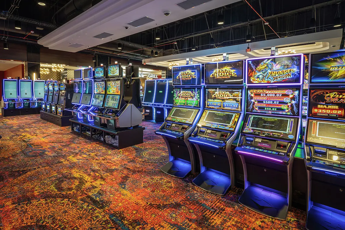 A vibrant casino interior with rows of illuminated slot machines, colorful patterned carpet, and a lively atmosphere.