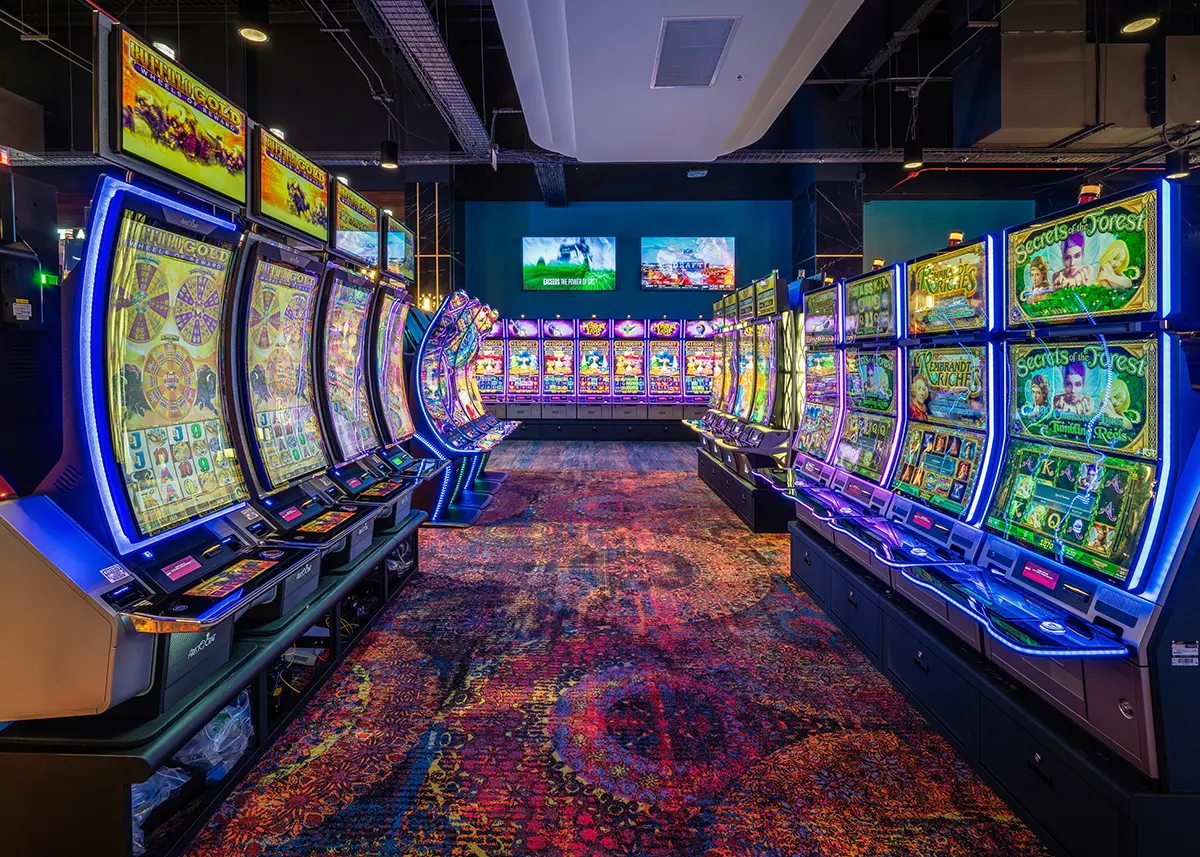 Bright casino interior featuring rows of colorful slot machines with vibrant screens. Decorative carpet adds to the lively, energetic feel.