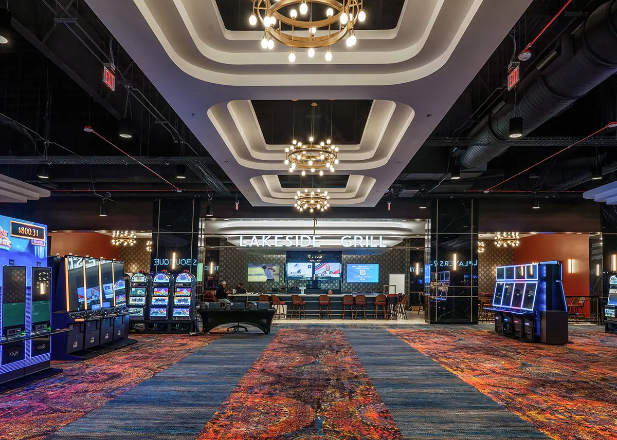 Casino interior with colorful carpet and modern chandeliers. Slot machines flank the path leading to the "Lakeside Grill," where TVs line the wall.