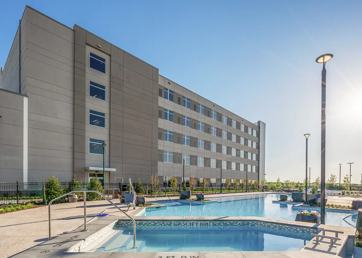 Modern hotel with a five-story facade beside a clear blue swimming pool under a sunny sky. Relaxing outdoor ambiance with lush greenery.