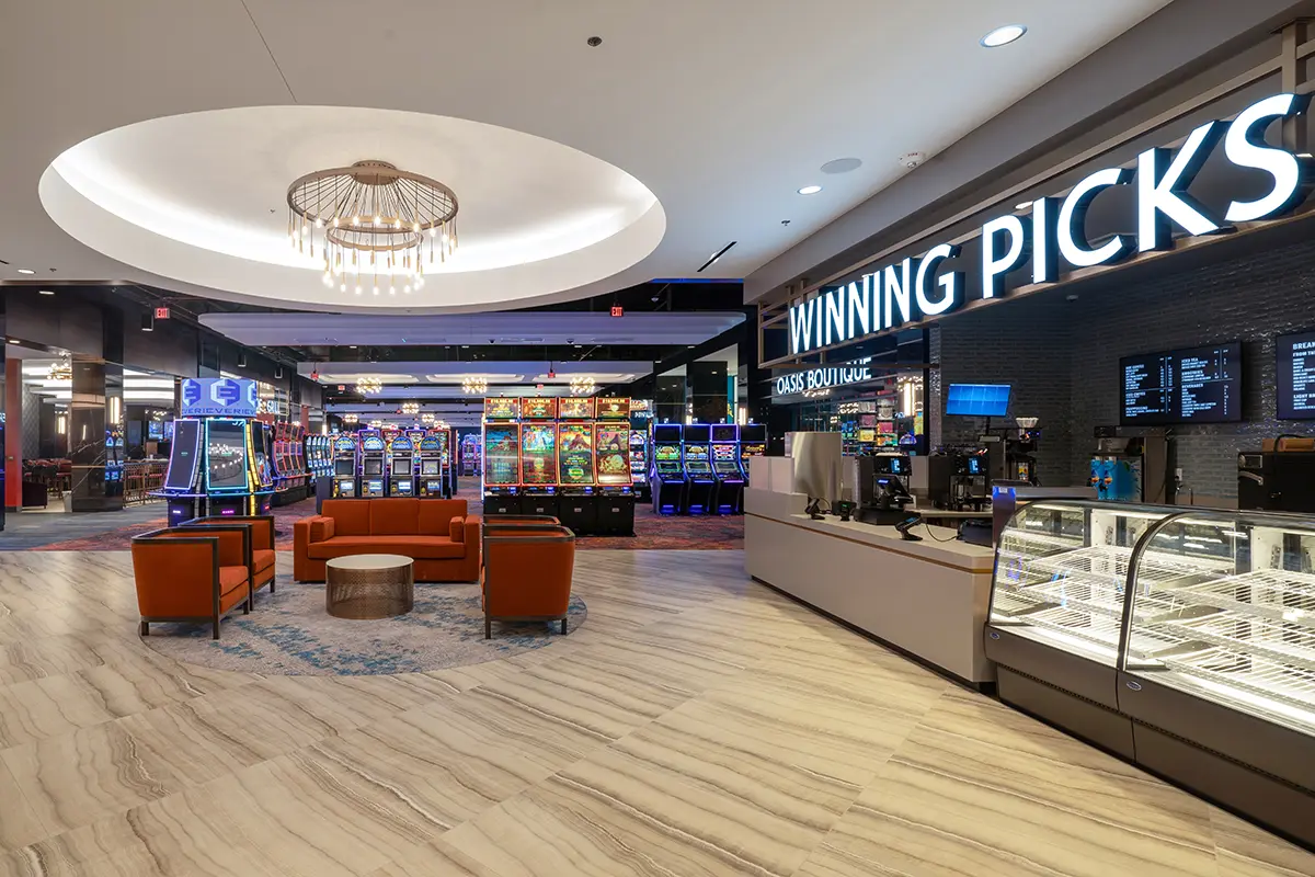 Modern casino interior featuring slot machines with colorful lights in the background. Foreground includes a seating area and an empty dessert display, under a bright chandelier. Signs read "Winning Picks."