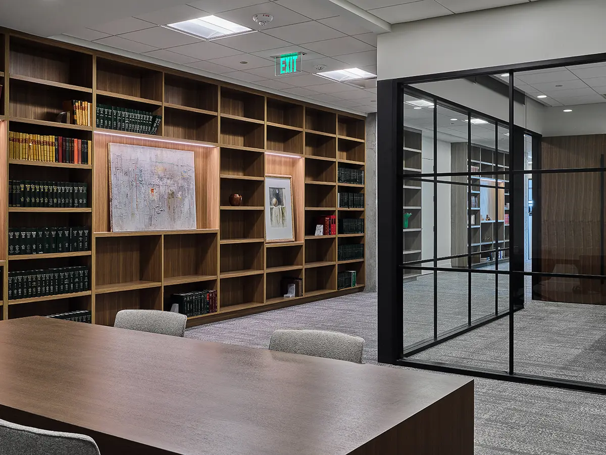 Modern office library with wooden shelves filled with books, framed art, and decor. Glass partition separates the room, creating an open, professional atmosphere.
