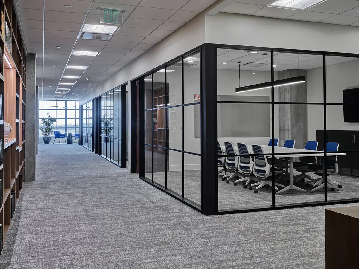 Modern office interior with glass-walled conference room on the right featuring a long table and blue chairs. Hallway on the left leads to windows. Calm ambiance.