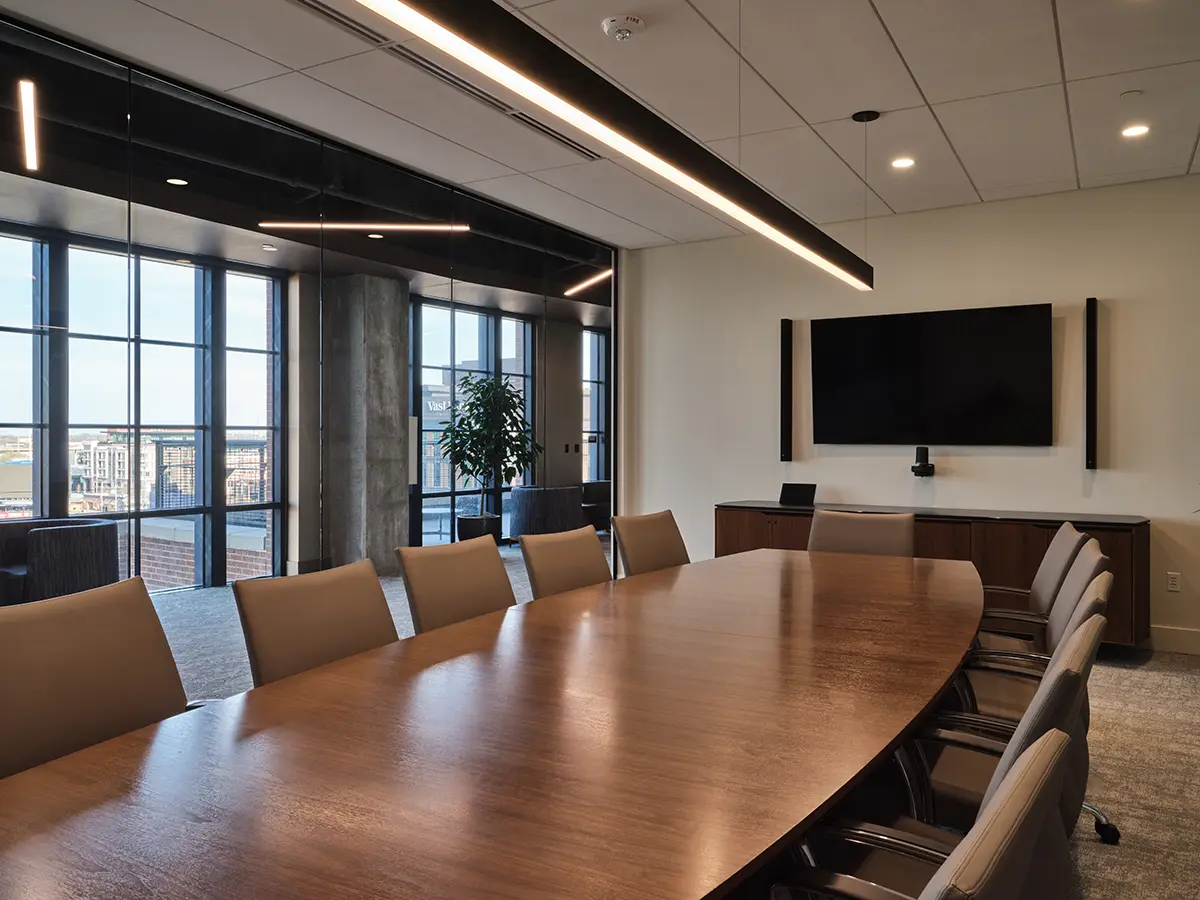 A modern conference room with a large oval wooden table, surrounded by beige chairs. A flat-screen TV is mounted on the wall, and floor-to-ceiling windows offer a city view. The room is minimally decorated, conveying a professional and sleek atmosphere.