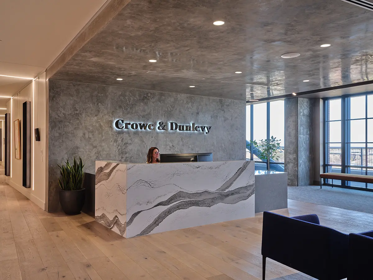 A modern conference room with a large oval wooden table, surrounded by beige chairs. A flat-screen TV is mounted on the wall, and floor-to-ceiling windows offer a city view. The room is minimally decorated, conveying a professional and sleek atmosphere.