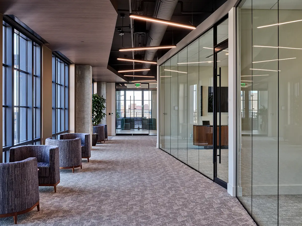 Modern office hallway with large windows and glass walls, featuring blue armchairs, carpeted floor, and sleek ceiling lights. Bright and professional atmosphere.