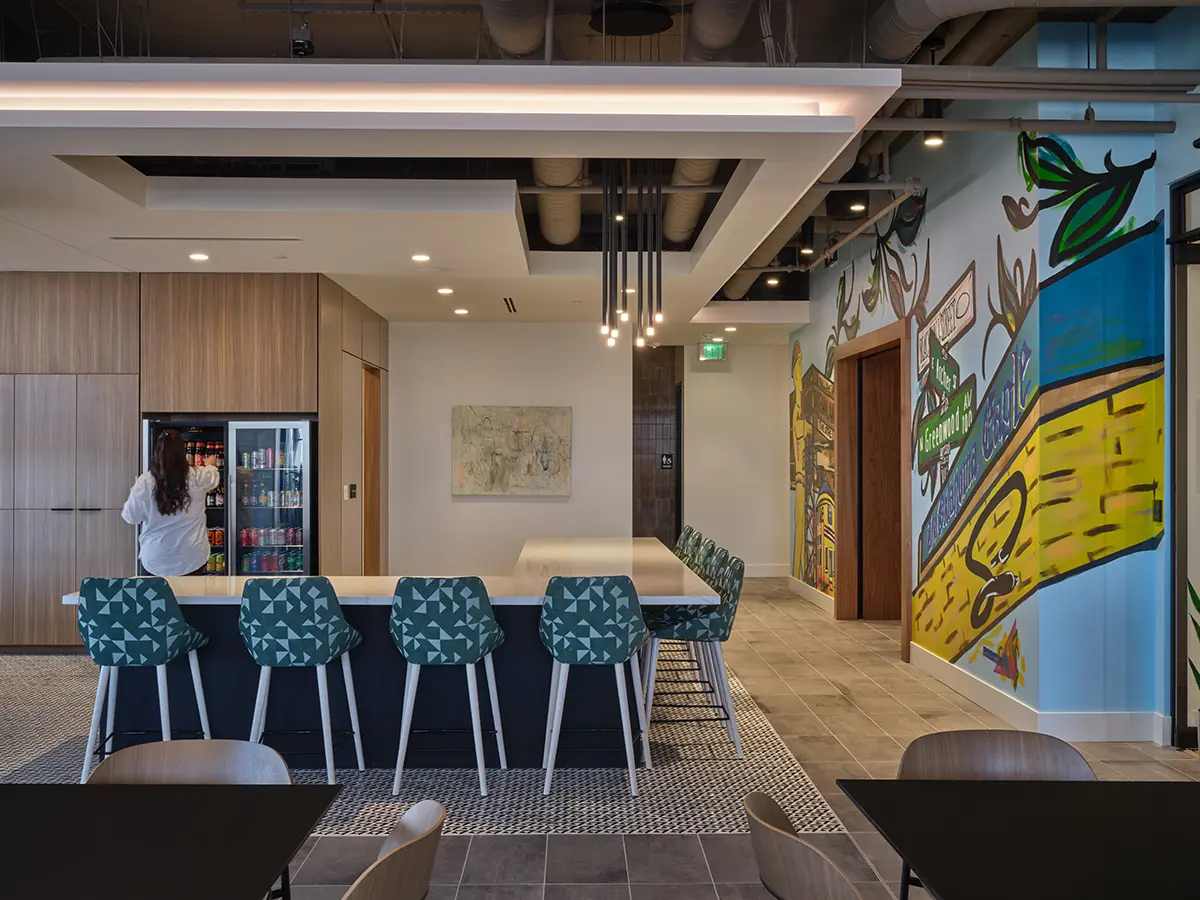 A modern office break room with a long bar counter, teal geometric-patterned stools, vibrant mural art, pendant lights, and a person accessing a beverage fridge.