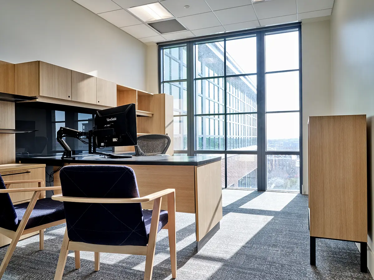Modern office with wooden furniture, computer on desk, and large windows letting in natural light. Two blue chairs face the desk, creating a welcoming atmosphere.