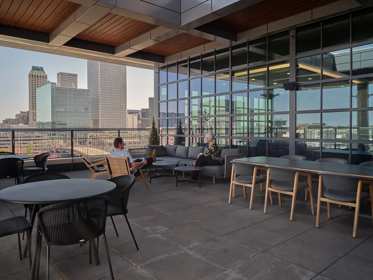 Outdoor rooftop patio with two people seated on a gray corner sofa, overlooking a city skyline. The setting is relaxed, featuring tables and chairs.