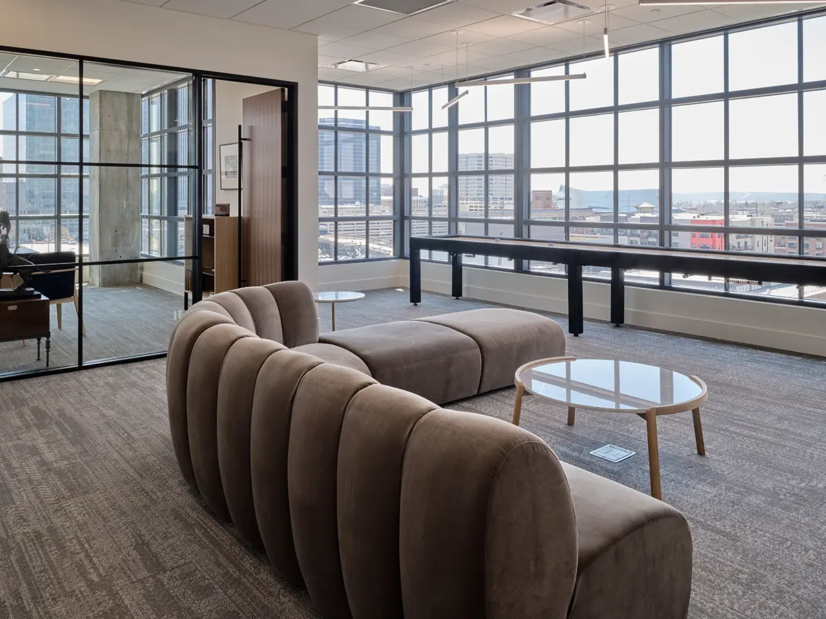Modern office lounge with curved gray couches and round glass tables. Large windows offer a cityscape view, creating an open, airy atmosphere.
