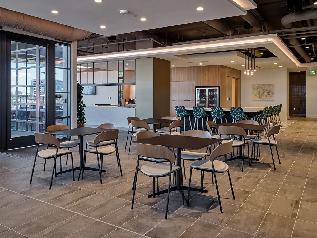 Modern cafeteria with wooden tables and chairs, tile flooring, a bar with green patterned stools, and pendant lighting. Bright and spacious ambiance.