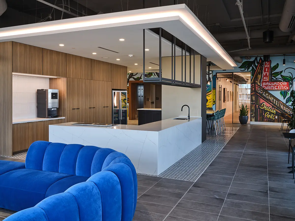 Modern office kitchen with a sleek white island, wood cabinetry, and a blue velvet couch. Bright mural in the background adds vibrant contrast.
