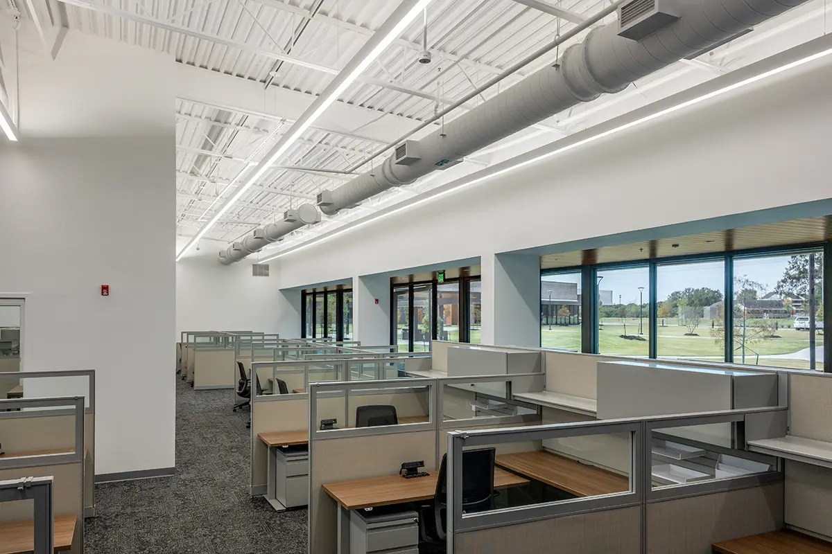 Modern office space with rows of empty cubicles, large windows letting in natural light, and an industrial ceiling design. The atmosphere is clean and open.