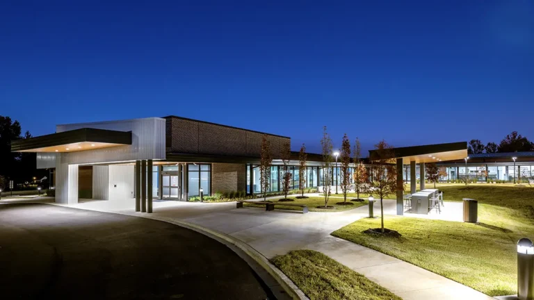 Modern building at dusk, with illuminated entrance and large windows, surrounded by grassy area and trees. The atmosphere is calm and inviting.