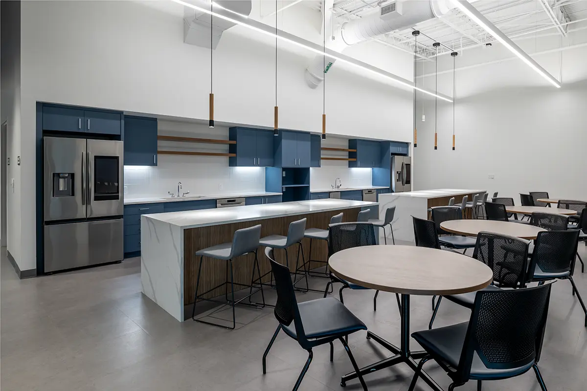 Modern office kitchen with blue cabinets, stainless steel appliances, and wooden round tables with chairs. Pendant lights hang from a high ceiling, creating a sleek, clean atmosphere.