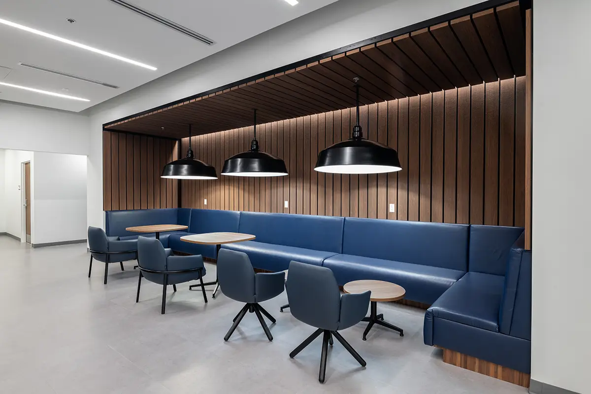 Modern lounge area with blue cushioned benches and swivel chairs around round tables. Wooden slatted walls, black pendant lights. Minimalist and sleek.