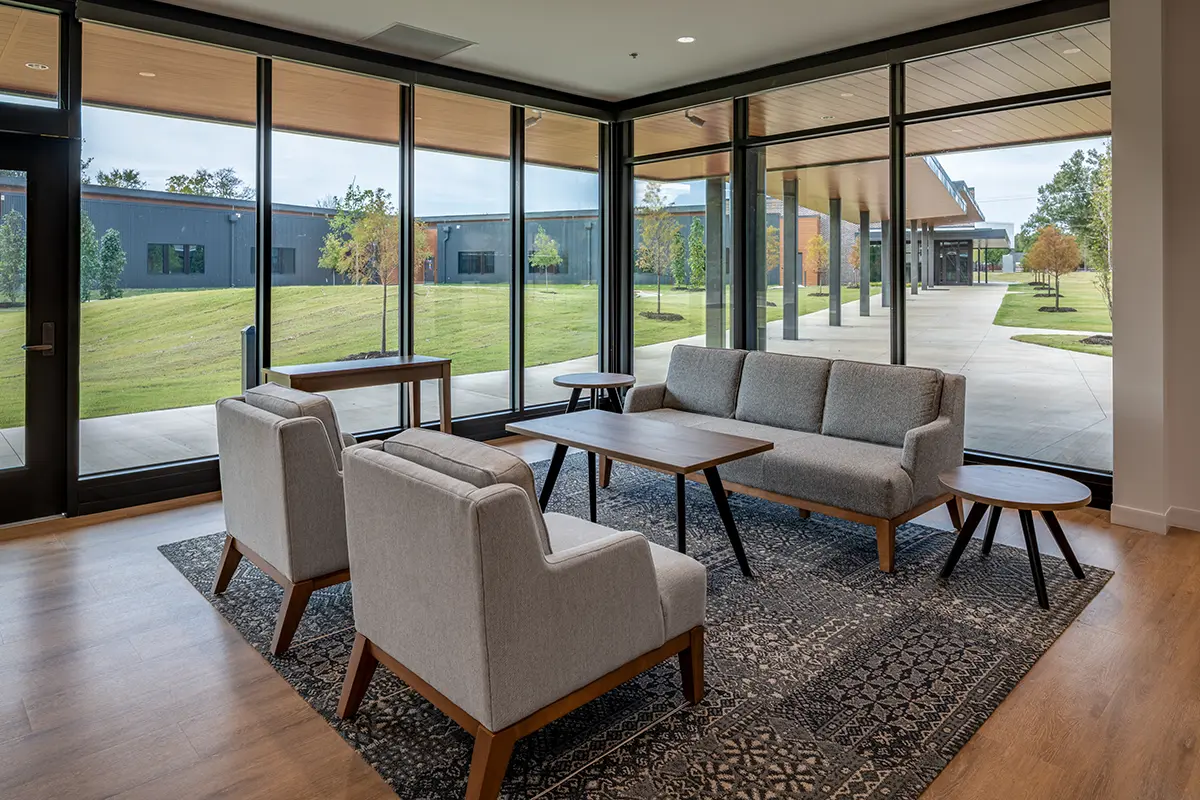 Modern office lounge with gray sofas, armchairs, and wooden tables on a patterned rug. Large windows offer a view of a green lawn and walkway. Cozy and inviting.