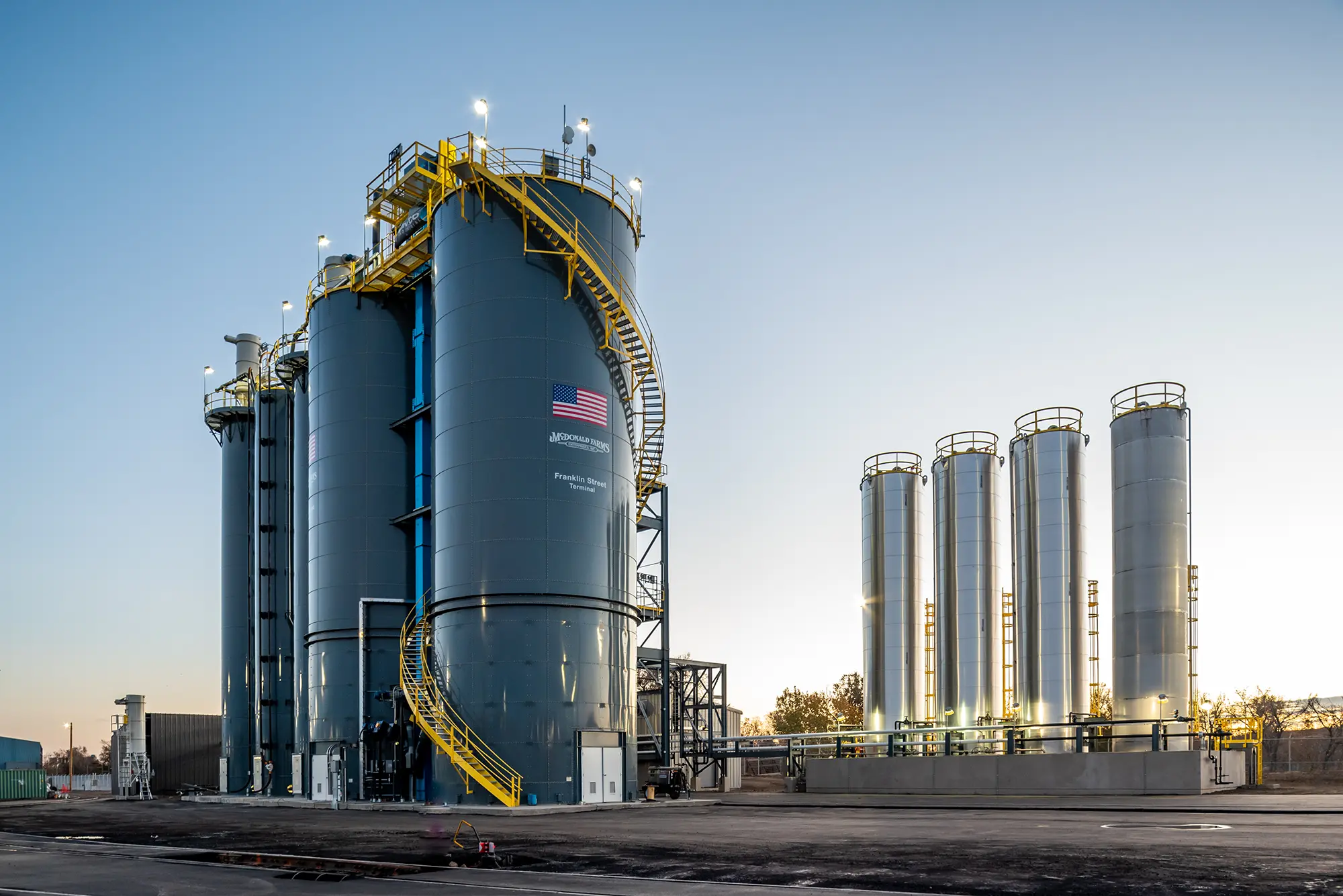 Large industrial silos stand at dawn with lights on, featuring a prominent American flag. The setting evokes a sense of progress and industry.