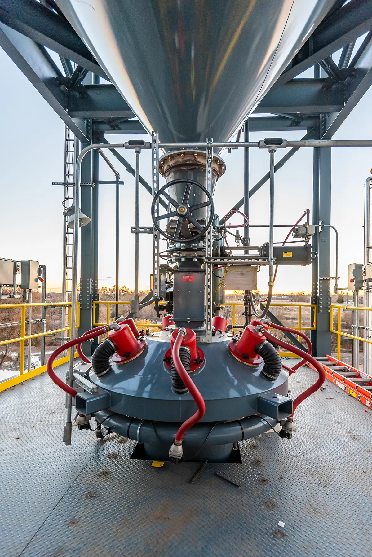 A large industrial machine with metal pipes and red hoses on a steel platform. The setting is outdoors with a clear sky, emphasizing a mechanical focus.
