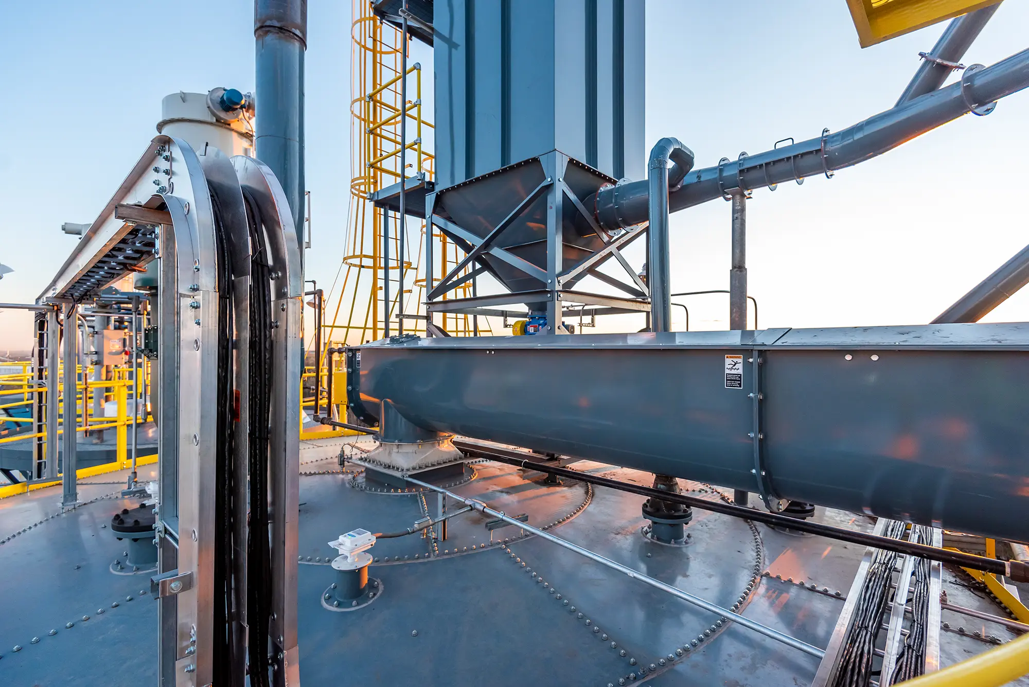 Industrial machinery with large metal structures, pipes, and yellow safety railings under a clear sky. The scene conveys efficiency and precision.