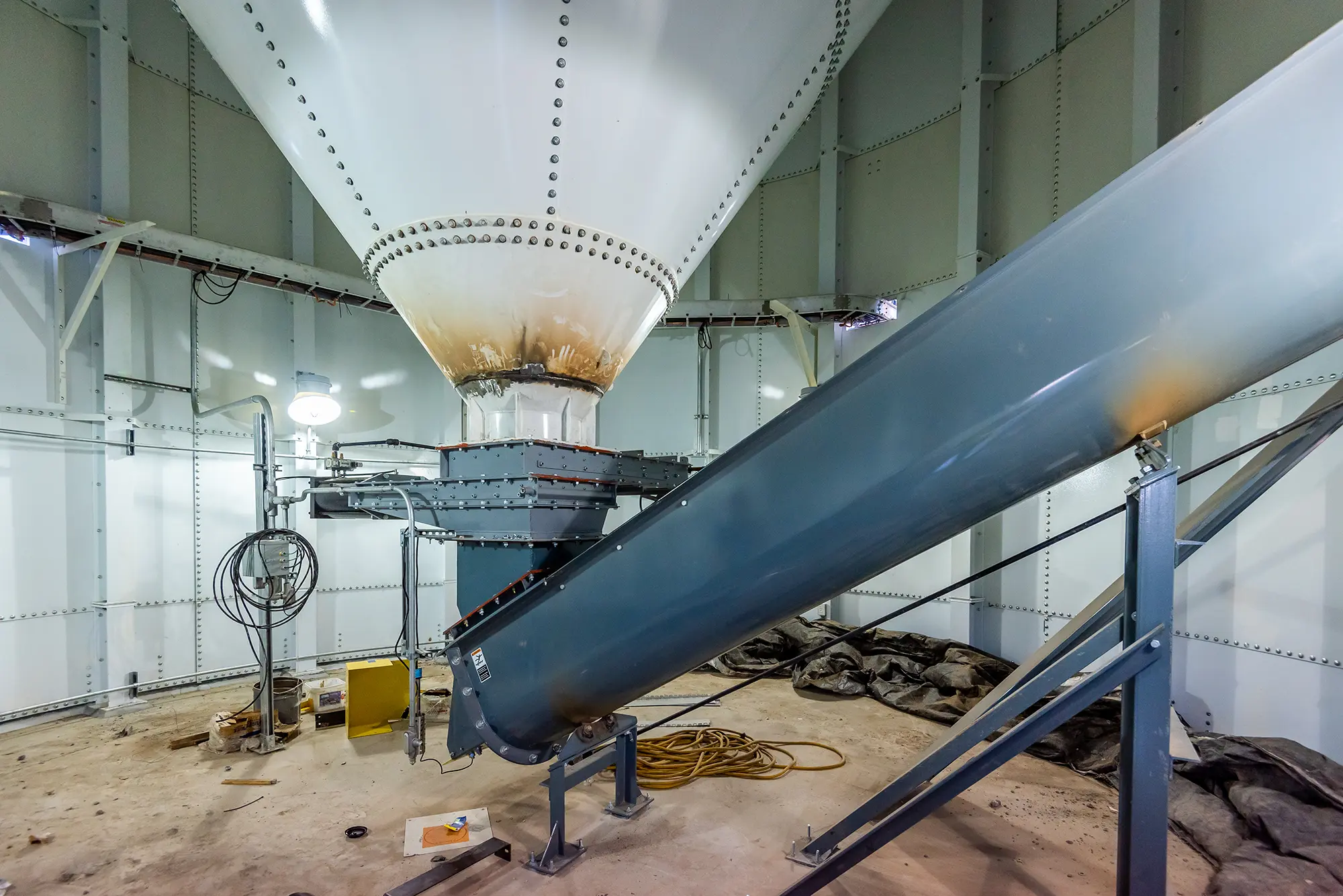 Industrial interior showing the bottom of a large white silo with a steel chute beside it. Cables, light, and scattered tools lie on a concrete floor.