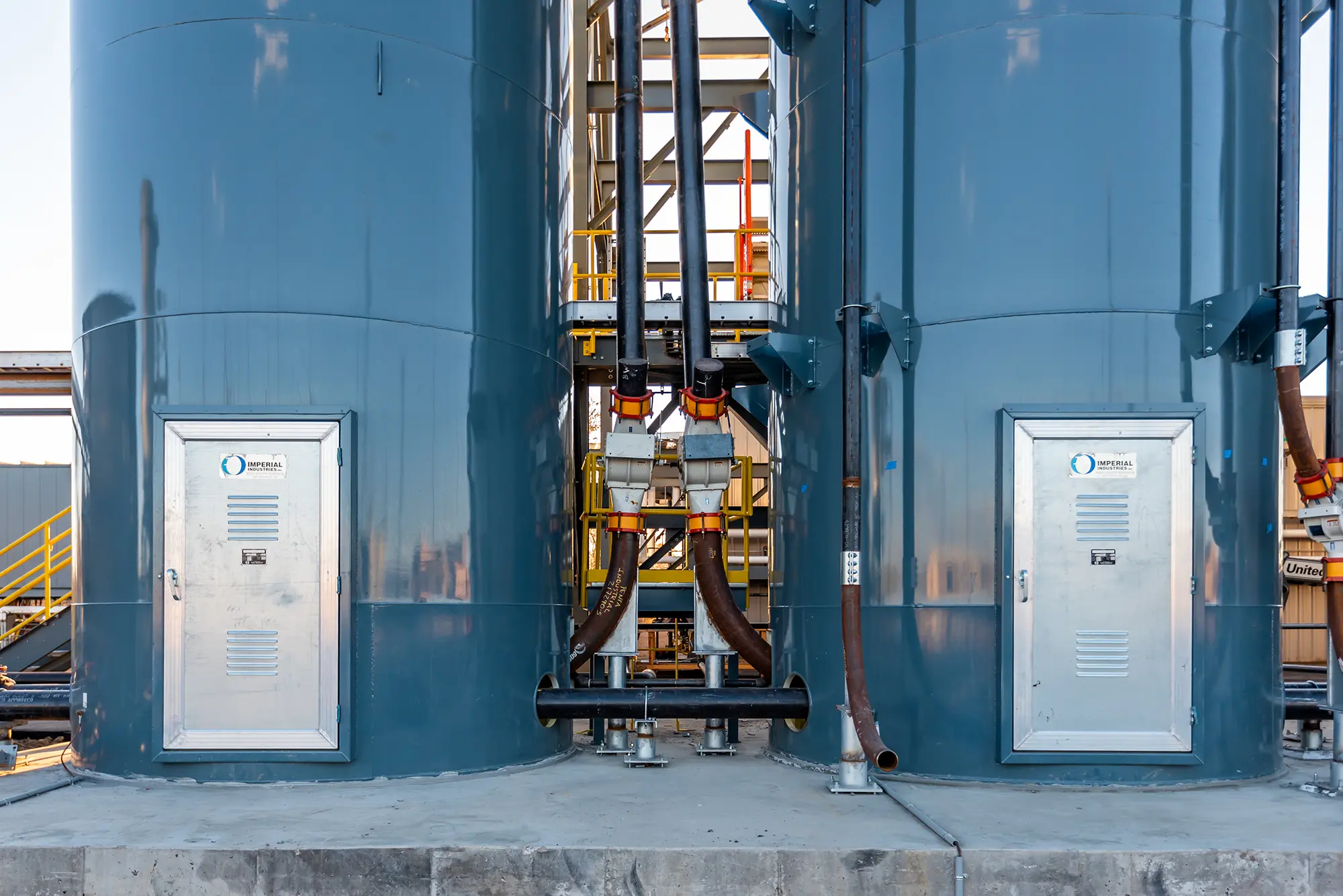 Two large blue industrial silos with metal doors stand side by side, connected by pipes. Bright yellow railings are visible in the background.