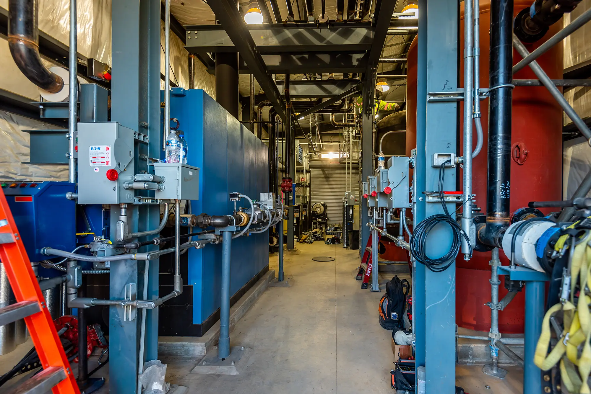 Industrial facility with complex machinery, including blue and red metal structures, cables, and pipes. The space is narrow and well-lit, conveying functionality.
