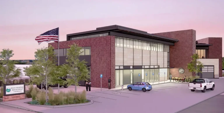 Modern two-story brick building with large windows, surrounded by trees and parked cars. An American flag flies near the entrance at dusk.