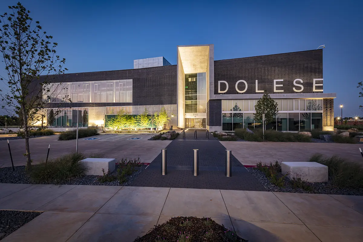 Modern two-story building with "DOLESE" sign, sleek architecture, large windows, and ambient evening lighting. Surrounded by neatly landscaped gardens.