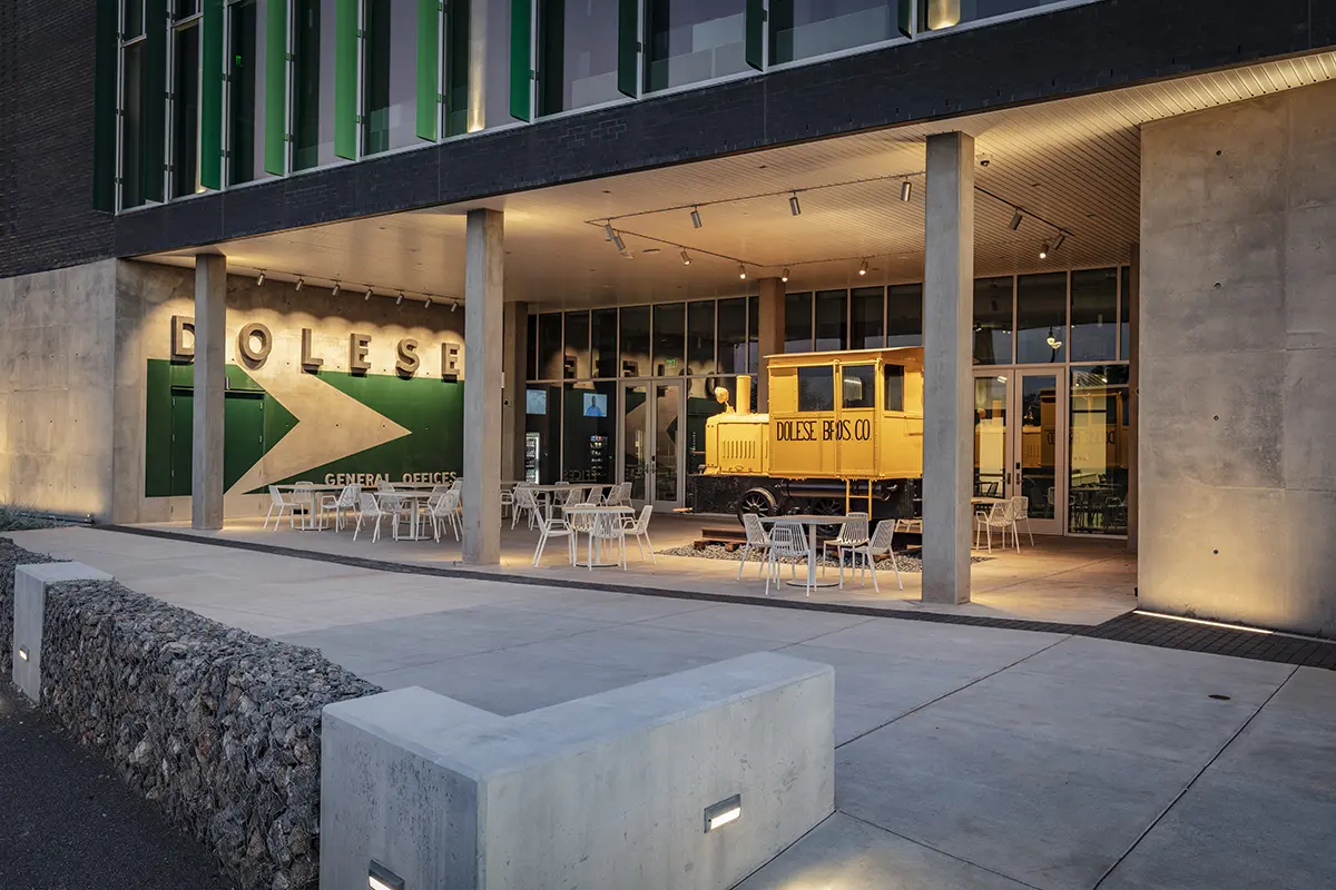 Modern building exterior at dusk with a well-lit entrance. Features include a yellow vintage truck labeled "DOLESE BROS CO" and outdoor seating.