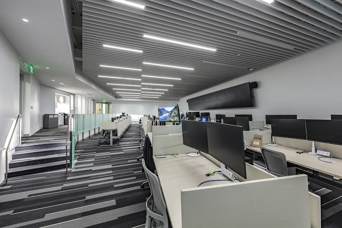 Modern, spacious office with multiple empty workstations and dual monitors. Sleek design, striped carpet, and a bright, professional atmosphere.