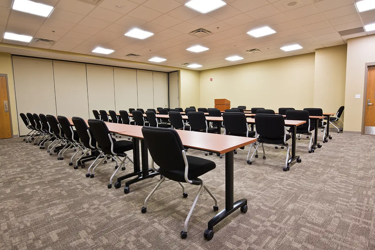 Training room with rows of black chairs and wooden tables facing a podium. The room has beige walls, carpeted floor, and a suspended ceiling with bright lights.